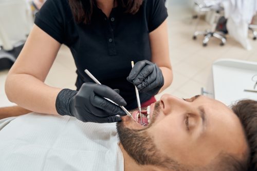 Closeup of male client lying on dental chair with opened mouth while doctor curing teeth. Professional dentist in uniform and protective gloves keeping probe and mirror and working. Concept of care.